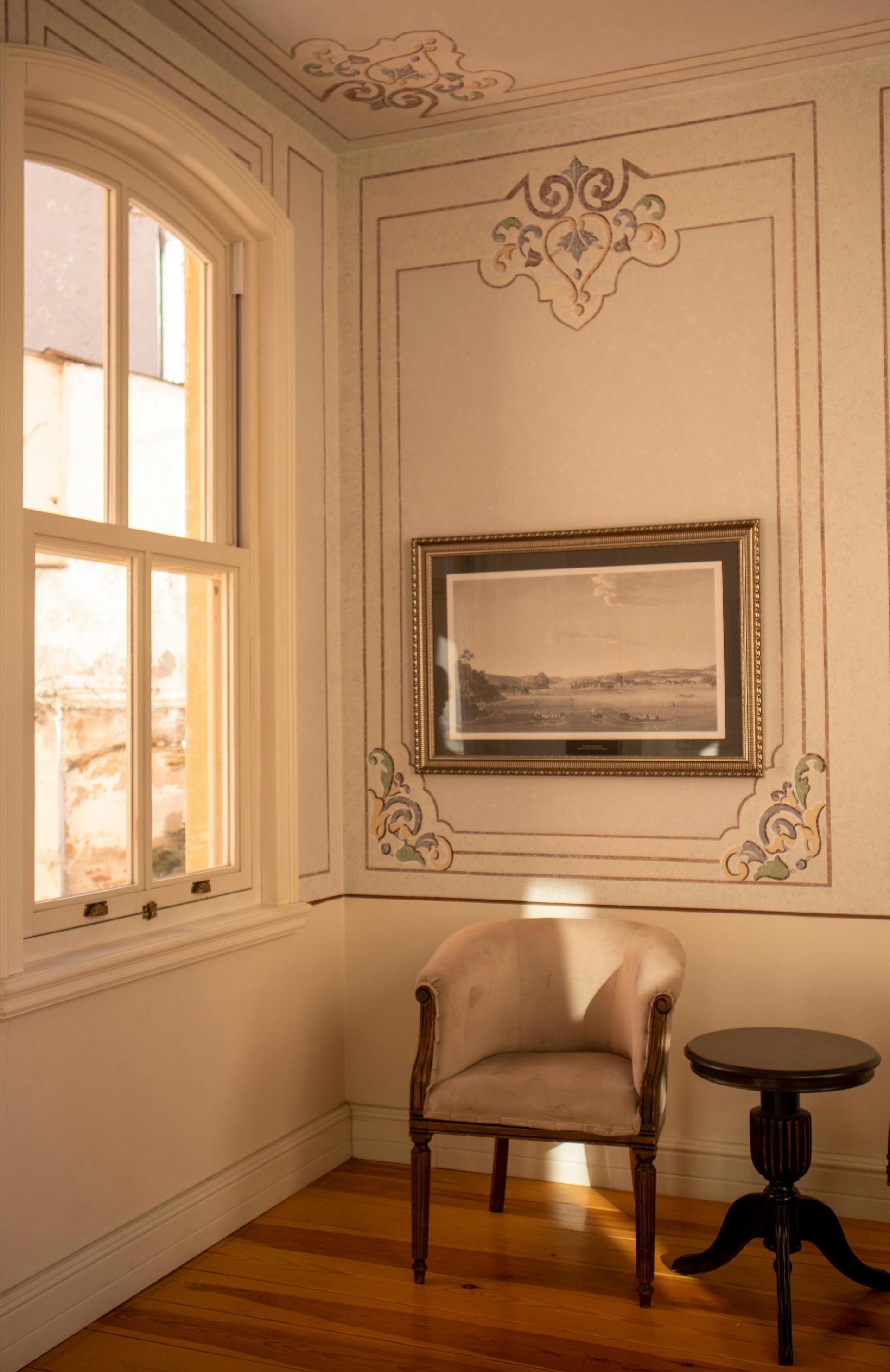 Corner of a room with a window, chair, and art. Sunlight streams in, illuminating the walls with ornate trim.