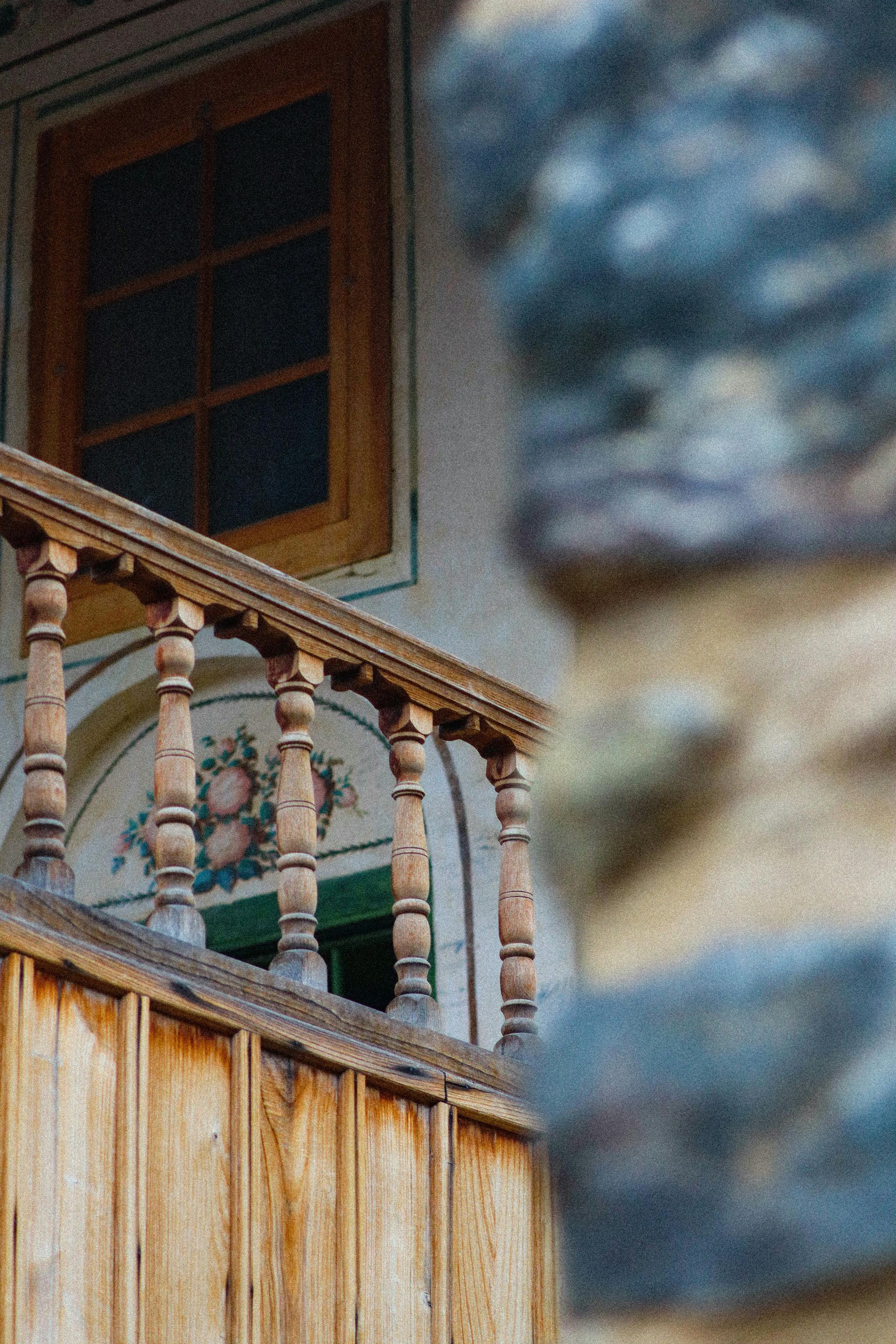 Balcony with wooden railing and window. Floral wall painting, aged wood, stone wall in foreground.