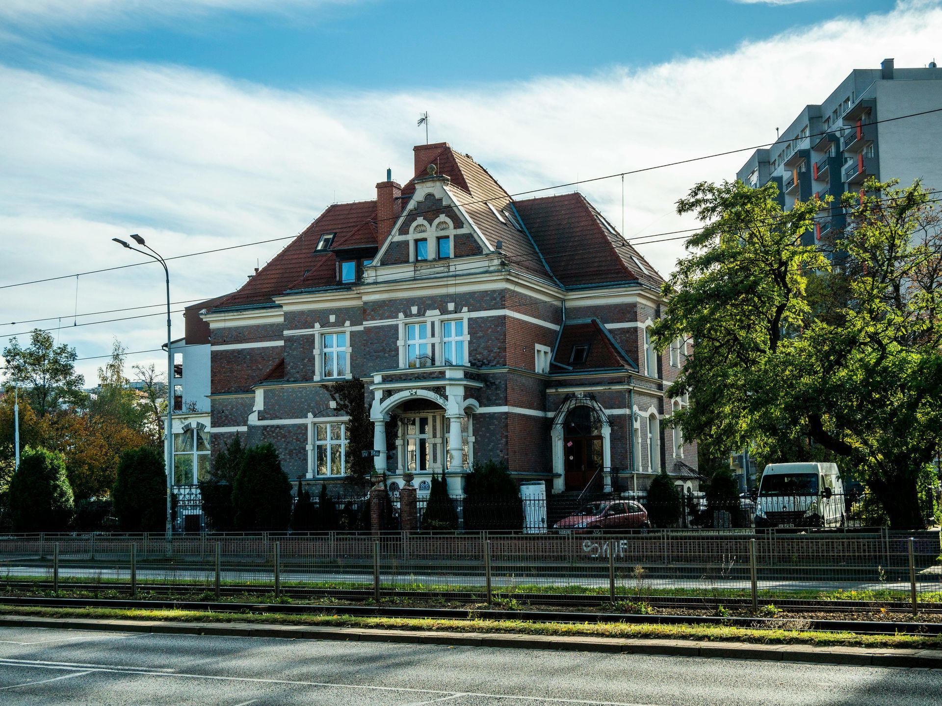 Brick house with red roof, ornate facade, set behind a metal fence. Green trees, blue sky in the background.