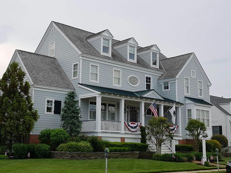Two-story light blue house with a porch and American flag, set in a well-manicured yard.