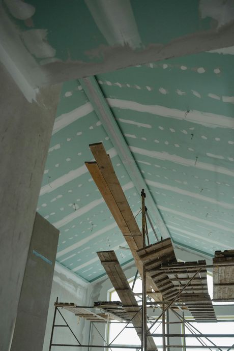 Construction site interior: drywall ceiling installation with scaffolding in place. Light green drywall, wood planks visible.