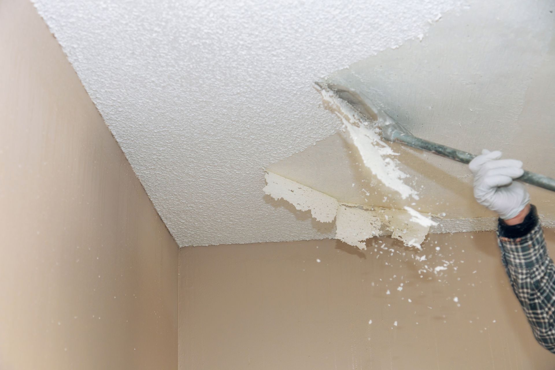 Person scraping textured ceiling with a wide scraper, flakes falling. Cream-colored walls.