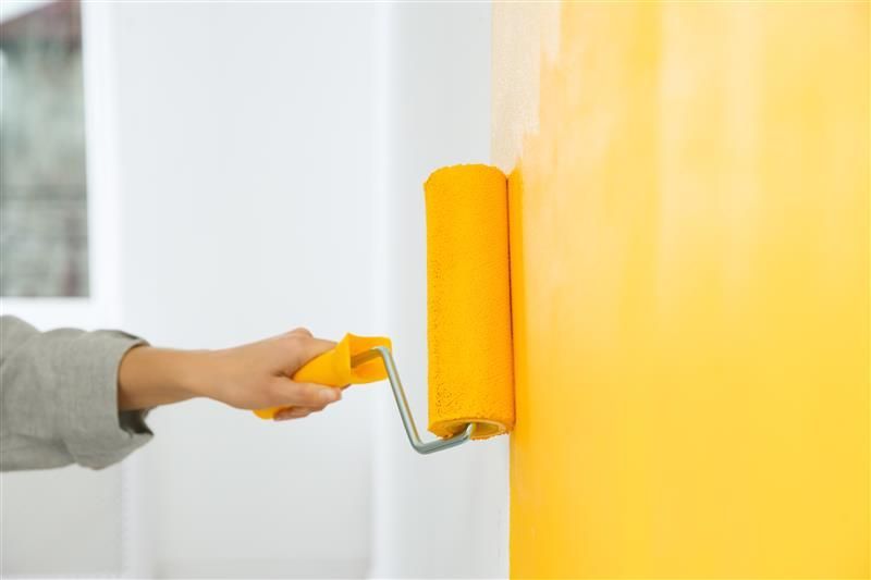 Hand holding a yellow paint roller, painting a yellow wall. White wall is adjacent.
