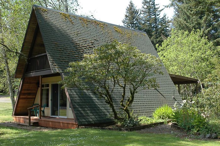 A small A-frame cabin with a steep shingled roof sits among trees and grass.
