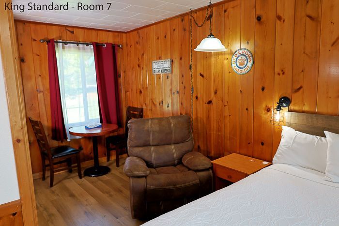 Cozy wood-paneled hotel room with bed, recliner, small table, lamp, and window with red curtains.