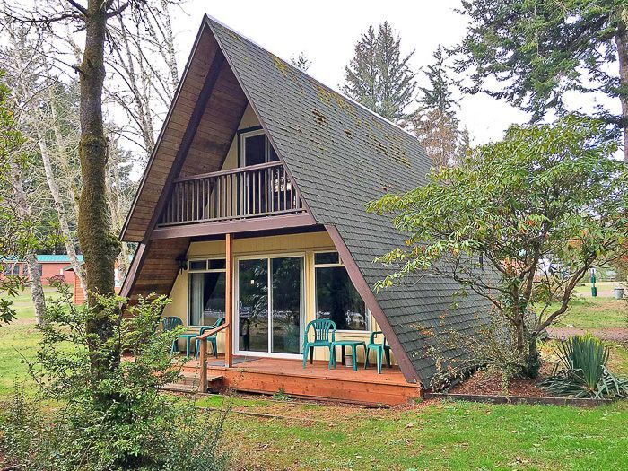 A-frame cabin with a steep roof, porch chairs, and a balcony nestled among trees and greenery.