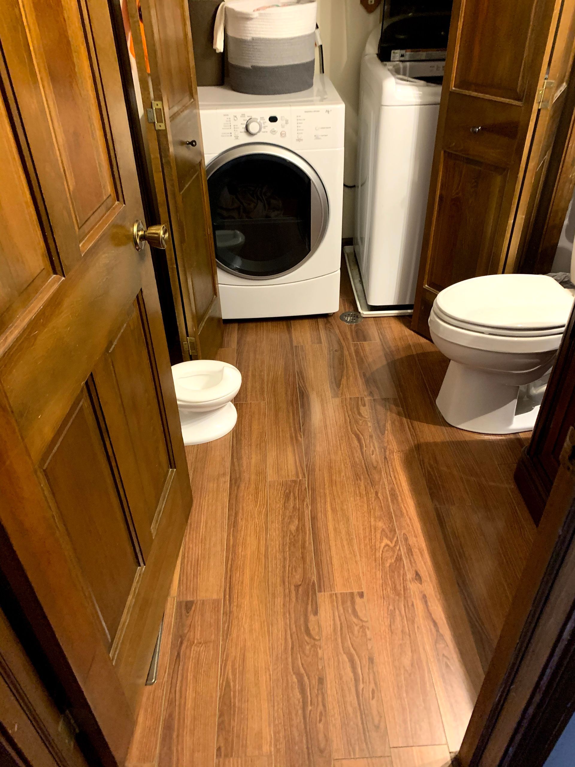 Narrow laundry room with a front-loading washer, dryer, toilet, and wooden floorboards.