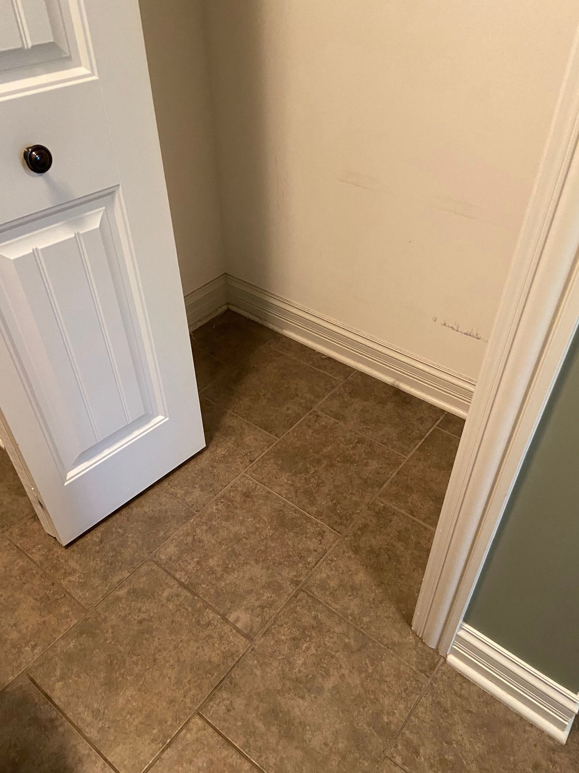 Small closet with open white door, beige walls, and brown tiled floor.