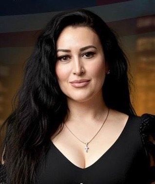 Woman with long black hair, wearing a black top and a cross necklace, smiling.