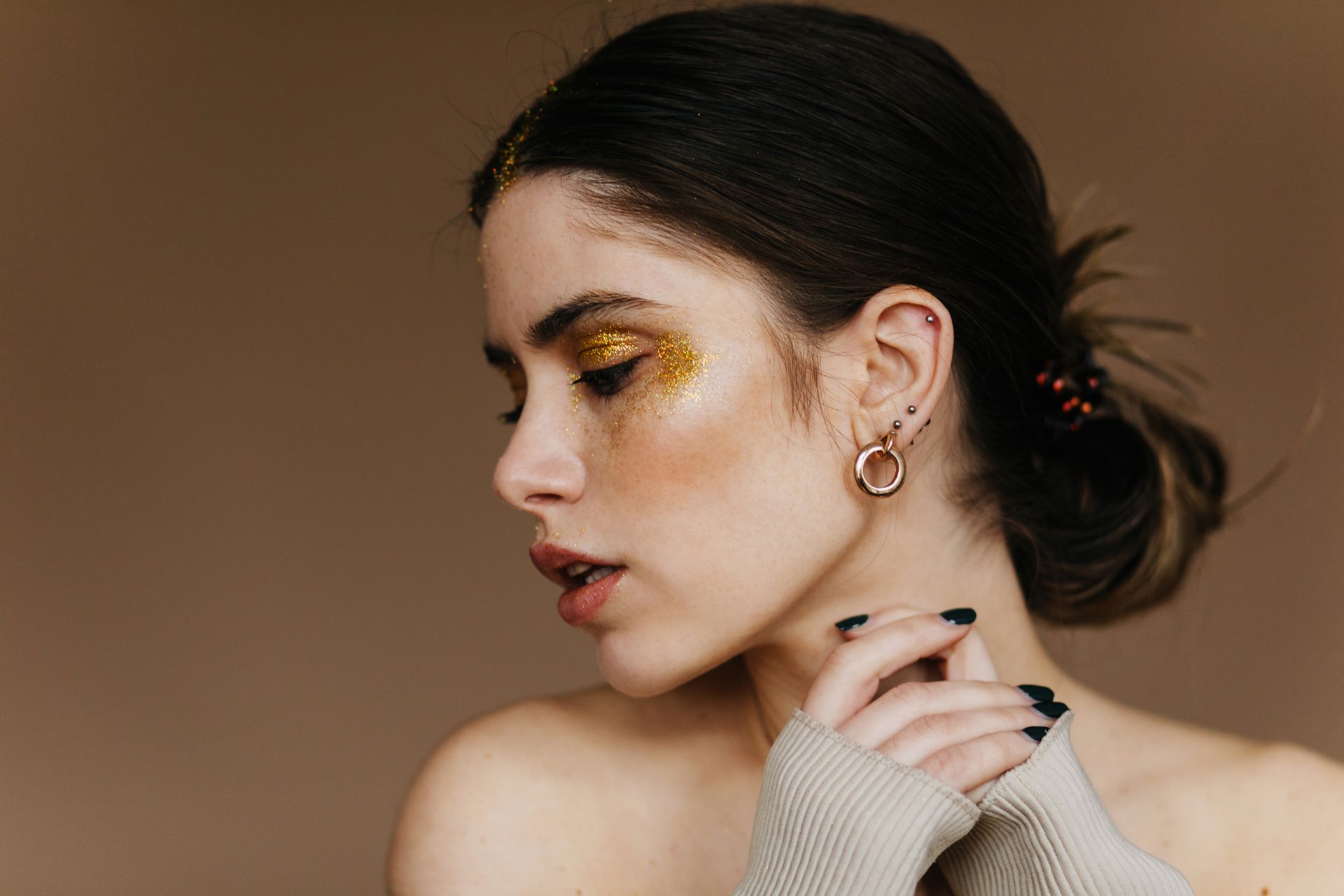 A woman with gold glitter on her face is wearing earrings and a sweater.