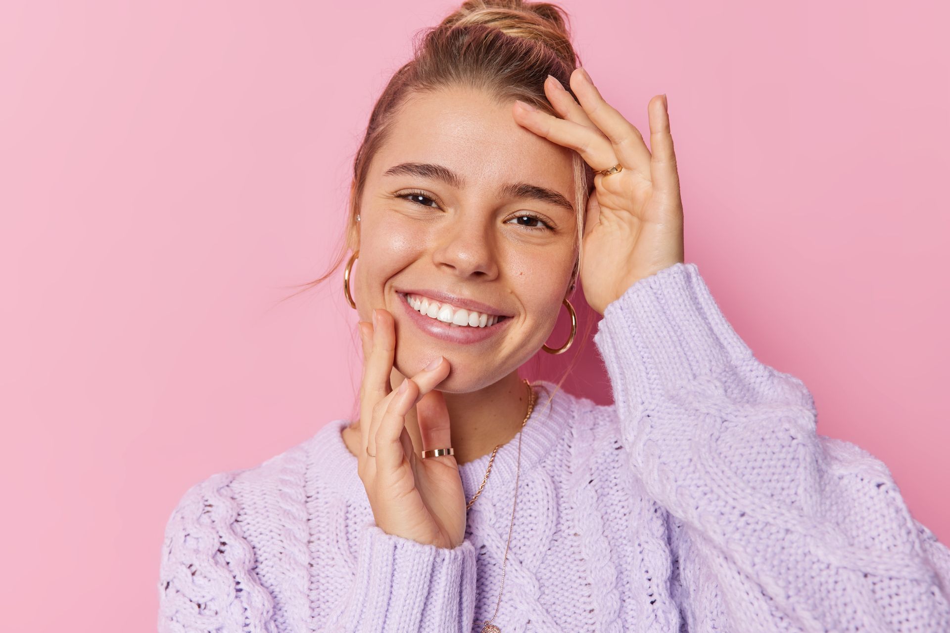 A woman in a purple sweater is smiling and touching her face.