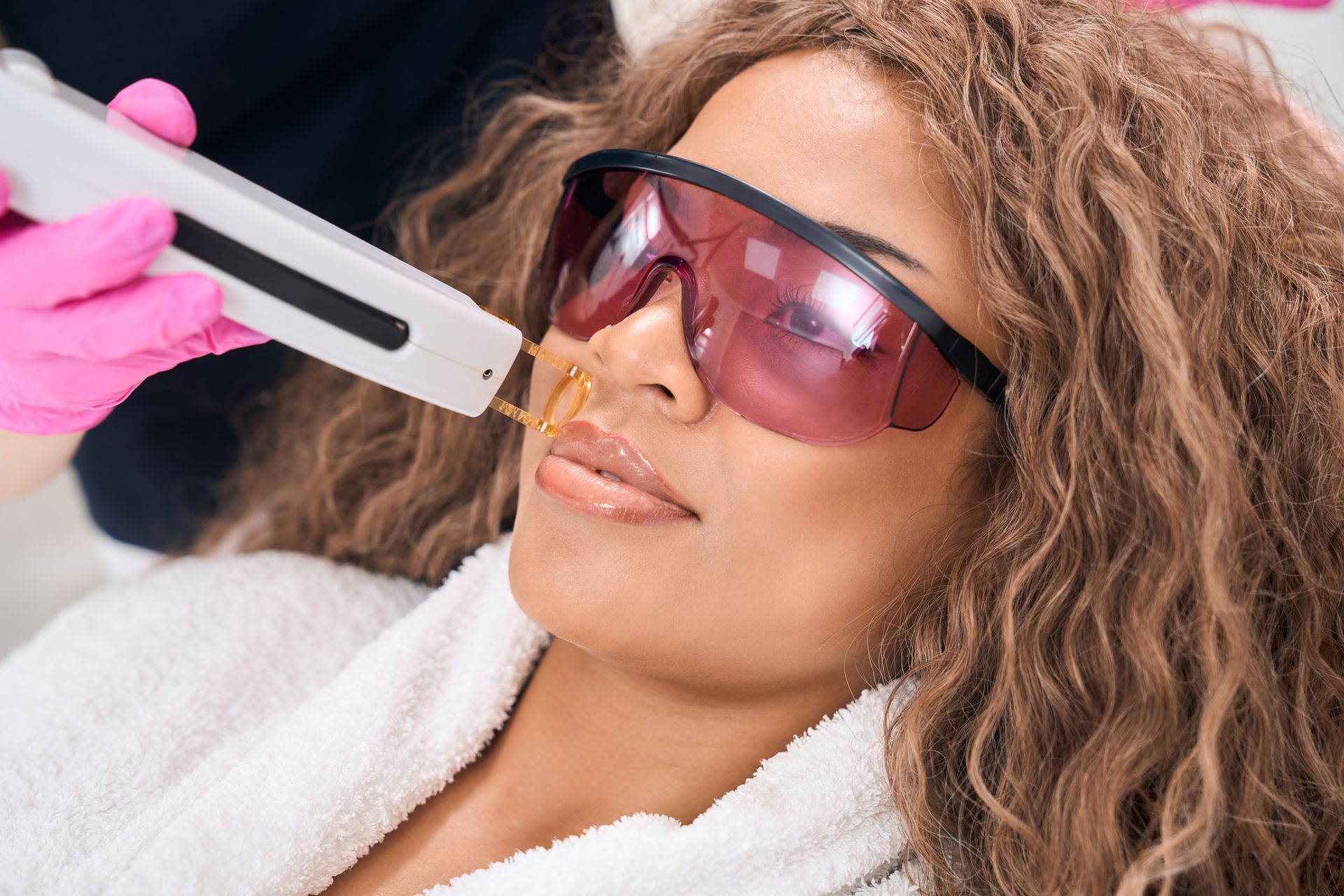Woman receiving laser hair removal on her upper lip, wearing protective eyewear; pink gloves.