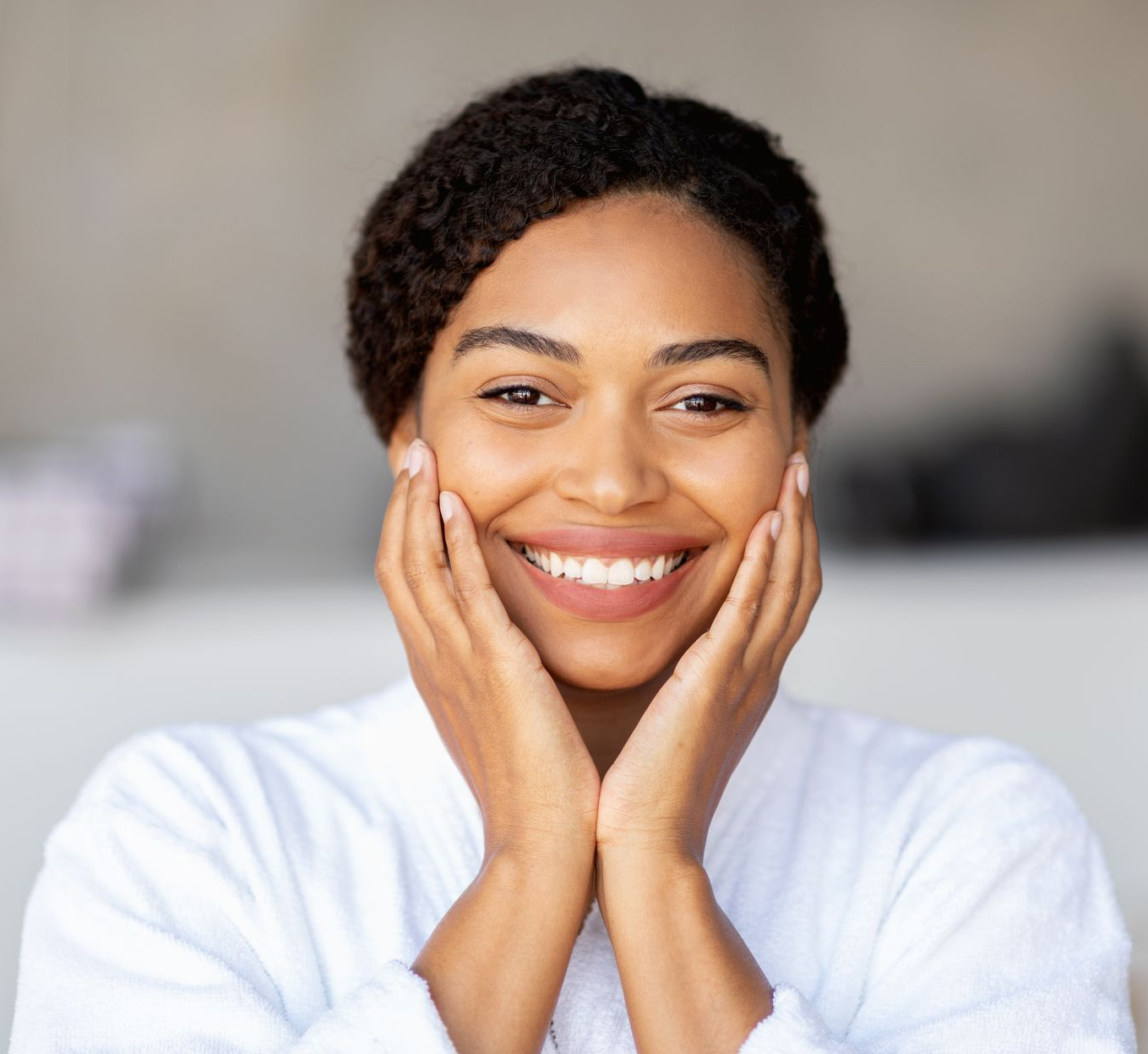 Woman smiling, holding hands on cheeks, wearing white robe.