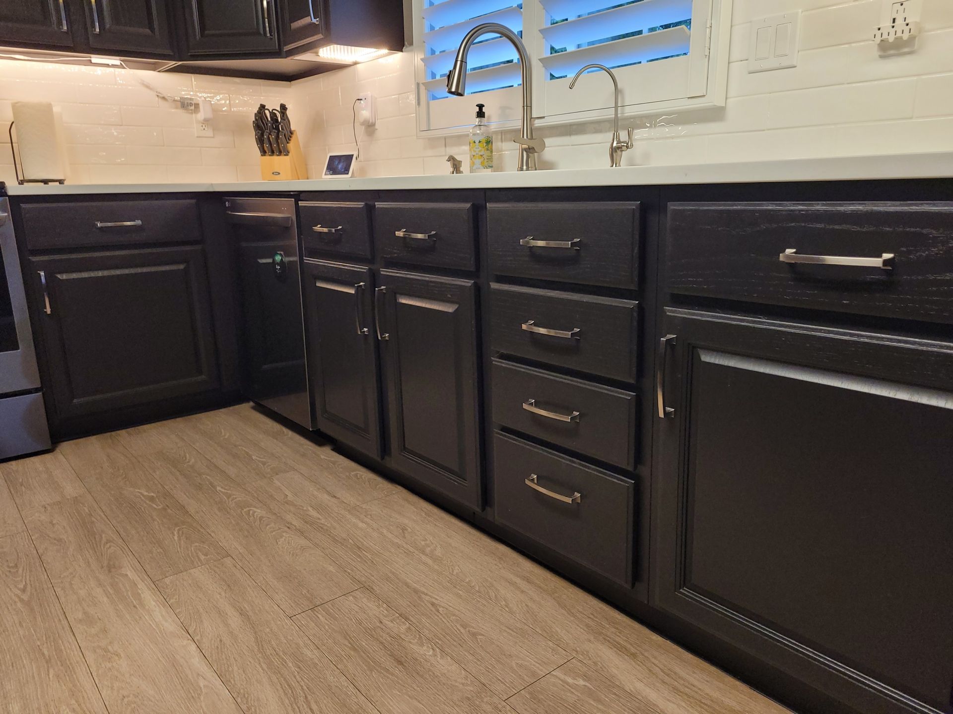 Black kitchen cabinets and wood floor beneath a white countertop and sink under a window with blinds