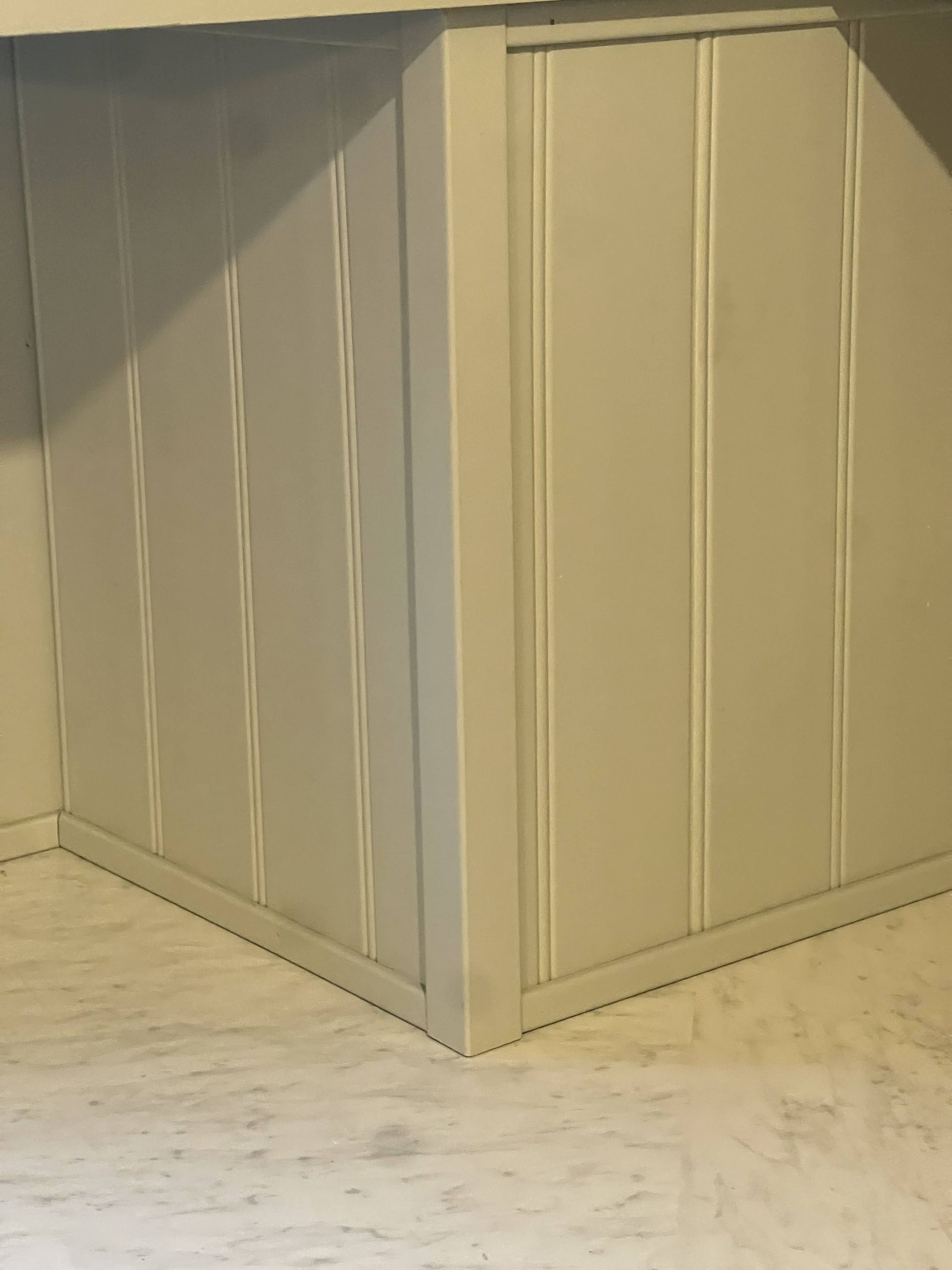 Corner of a white-painted wall with vertical paneling, meeting a light-colored floor.