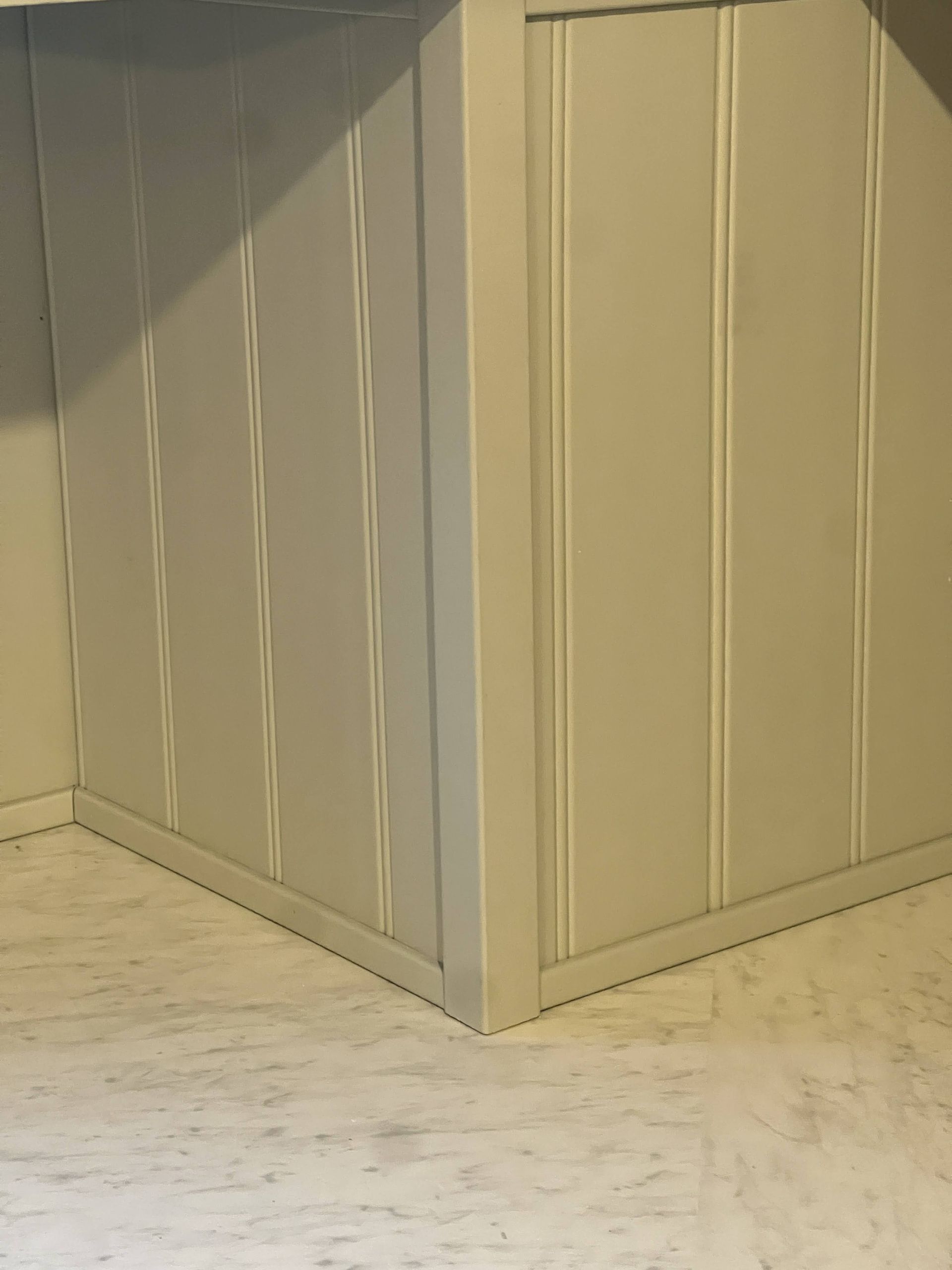 Corner of a room with light gray wainscoting and vertical paneling, meeting a light-colored floor.