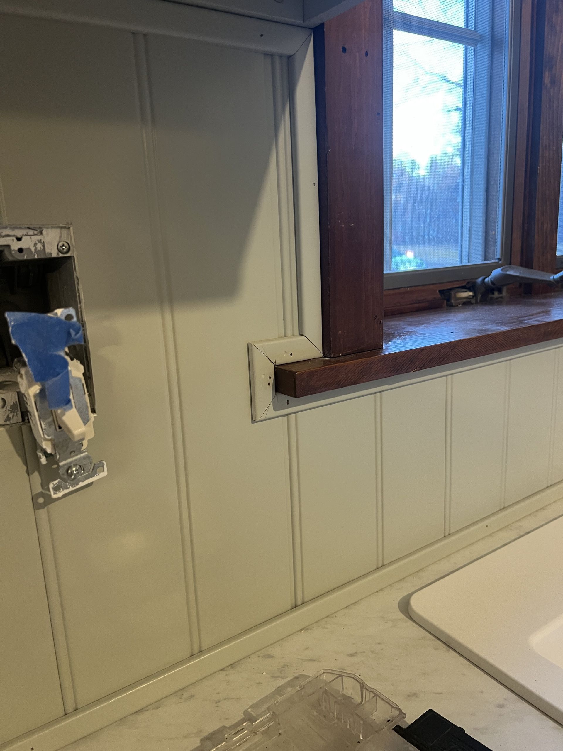 Kitchen wall with white paneling and a window with wooden trim. An electrical box is visible.