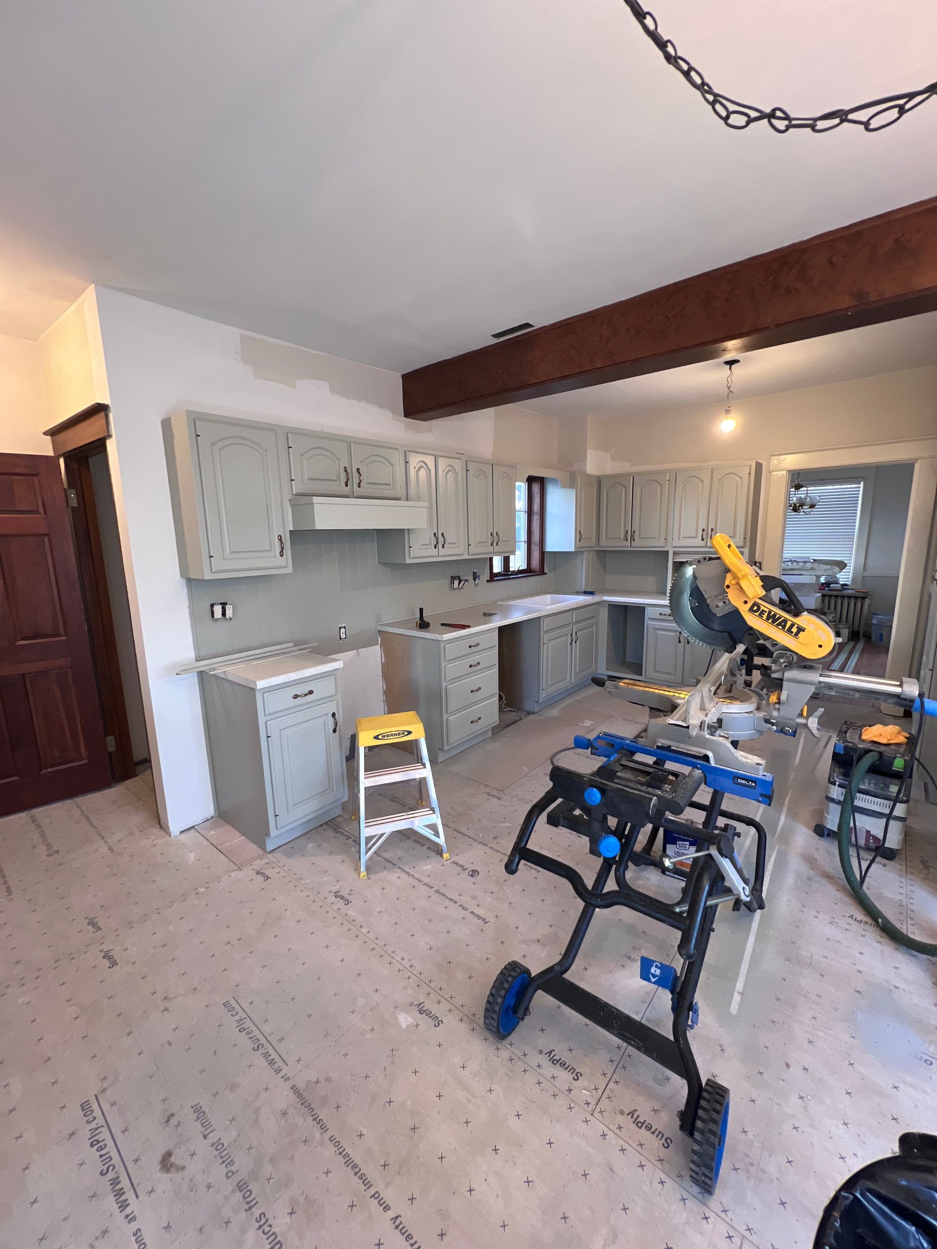 Kitchen under renovation with light gray cabinets, tools, and a wooden beam.