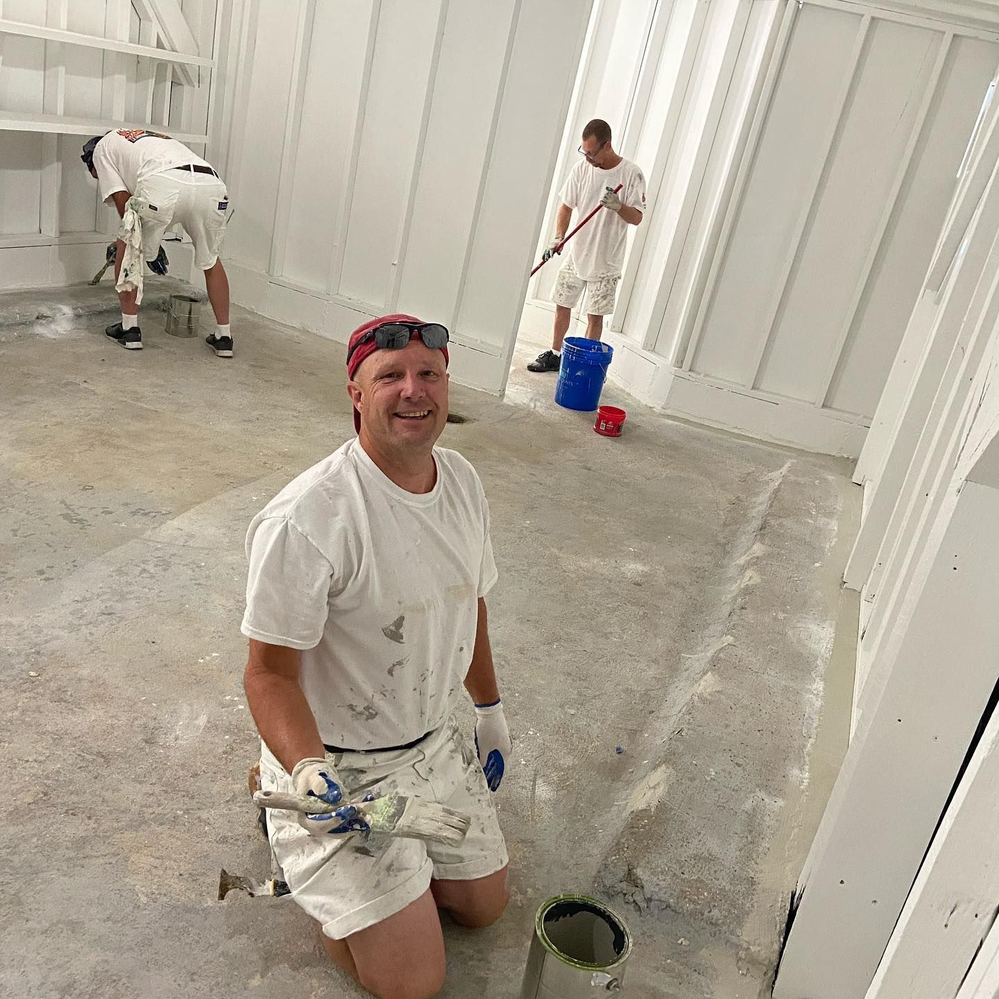 Three people paint a room white. One kneels, smiling, holding a brush. Others use brushes and rollers.