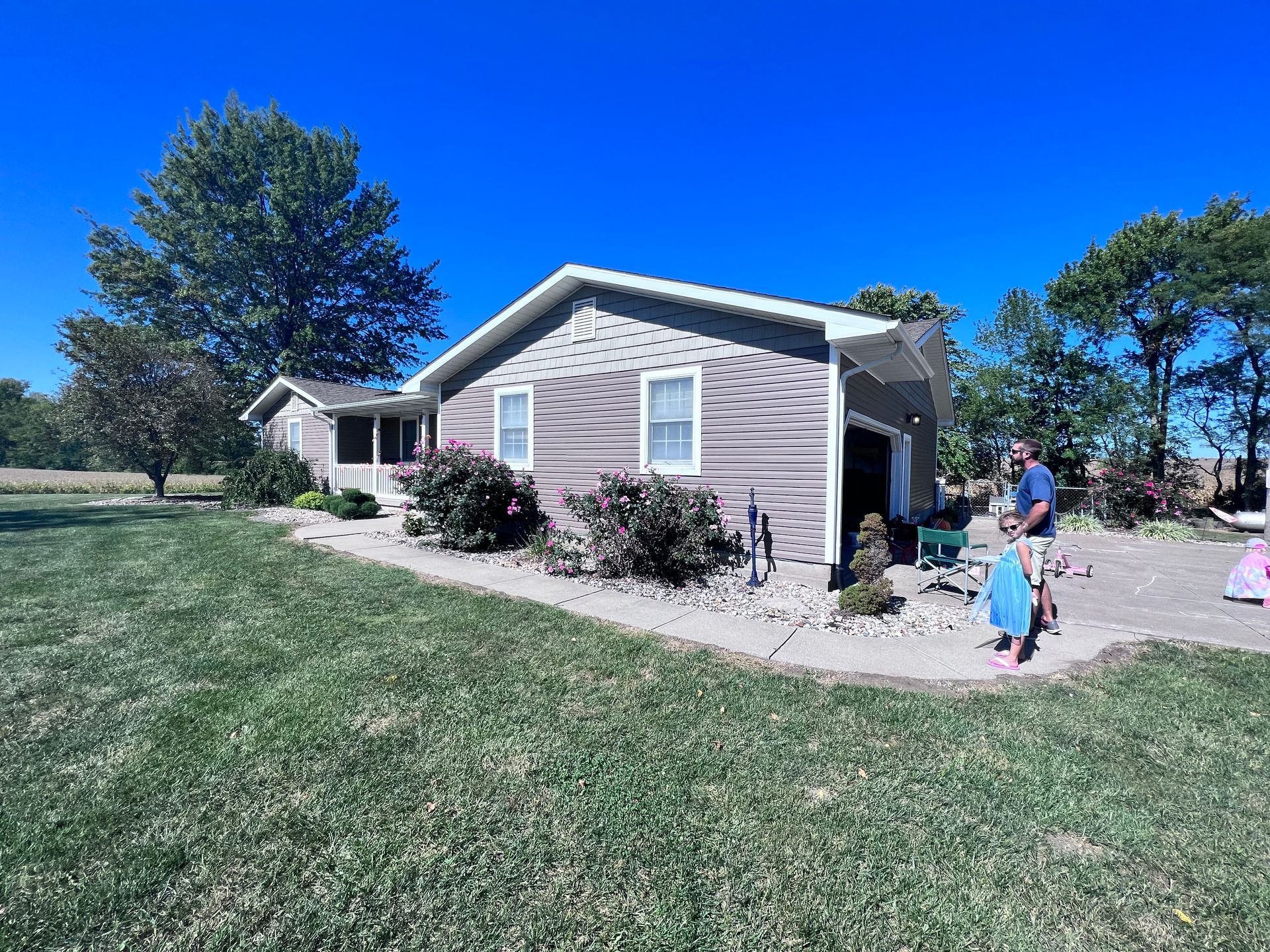 A low, brown house with a garage, blue sky, and a person with a child in the driveway.