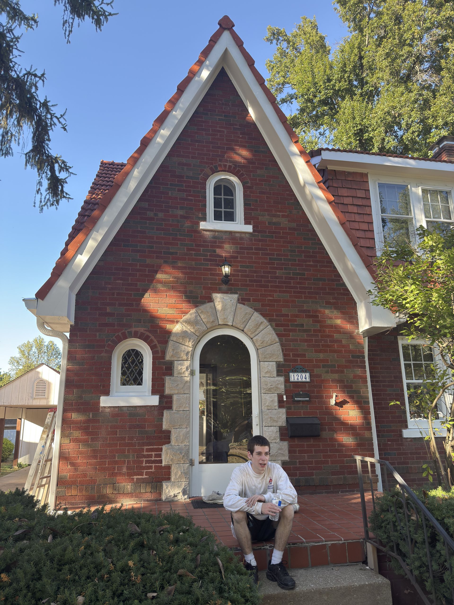 A person sits on steps of a red brick house with an arched doorway and a pointed roof under a blue sky.