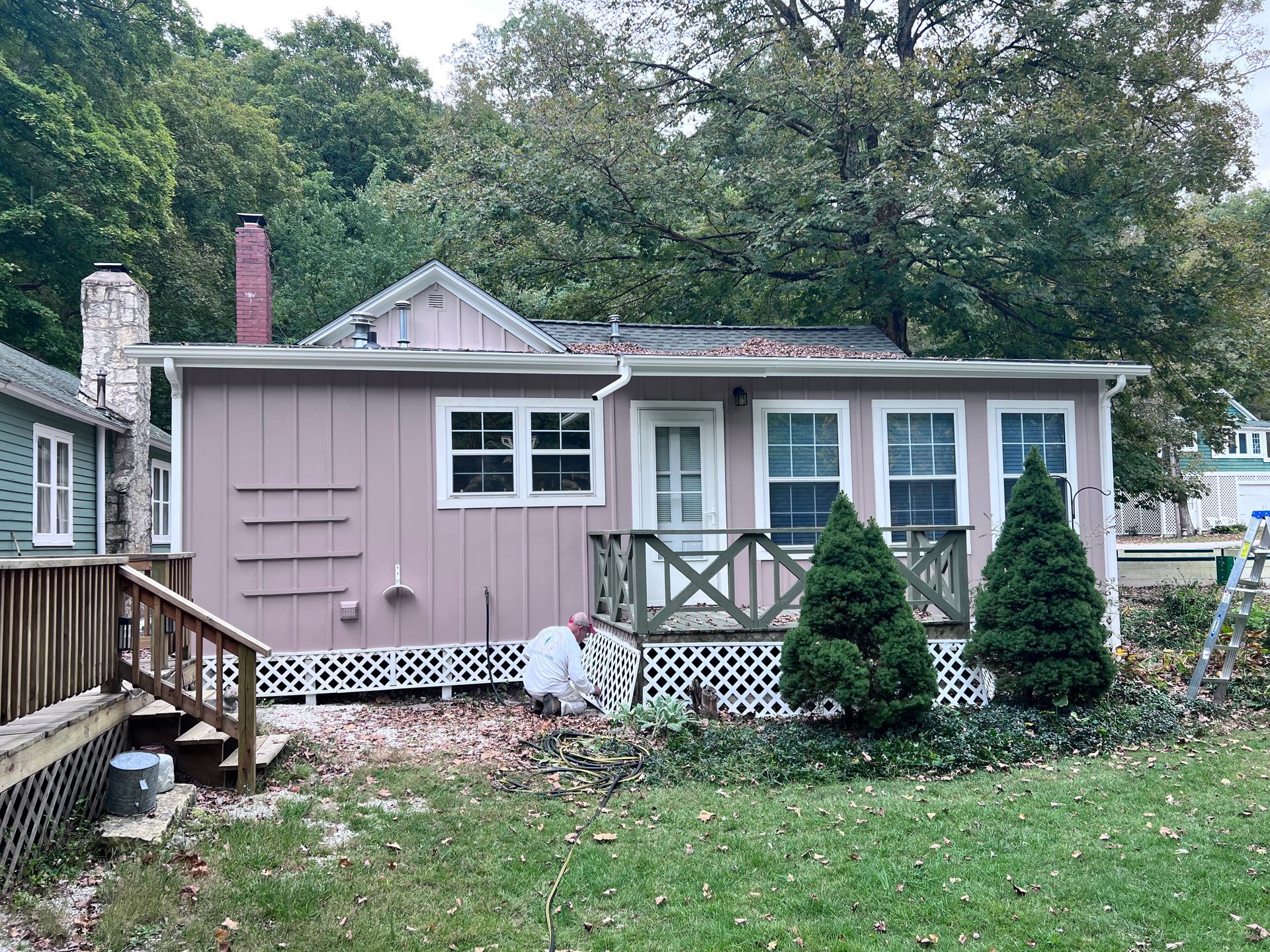 Small pink house with white trim, porch, and two evergreen bushes in front; fall foliage in the background.