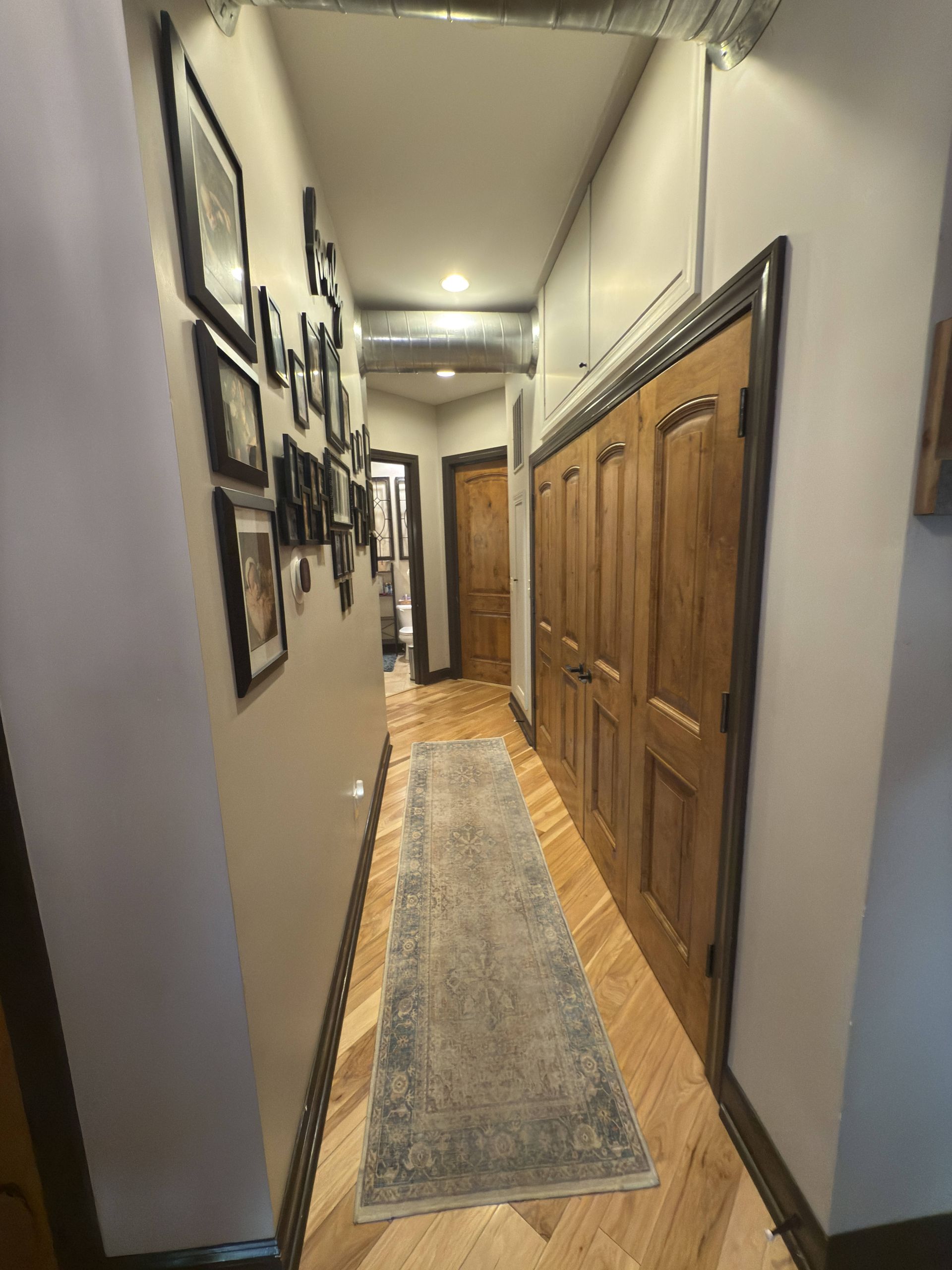 Narrow hallway with wooden floor, framed photos on the left, wooden closet doors on the right, and a rug.