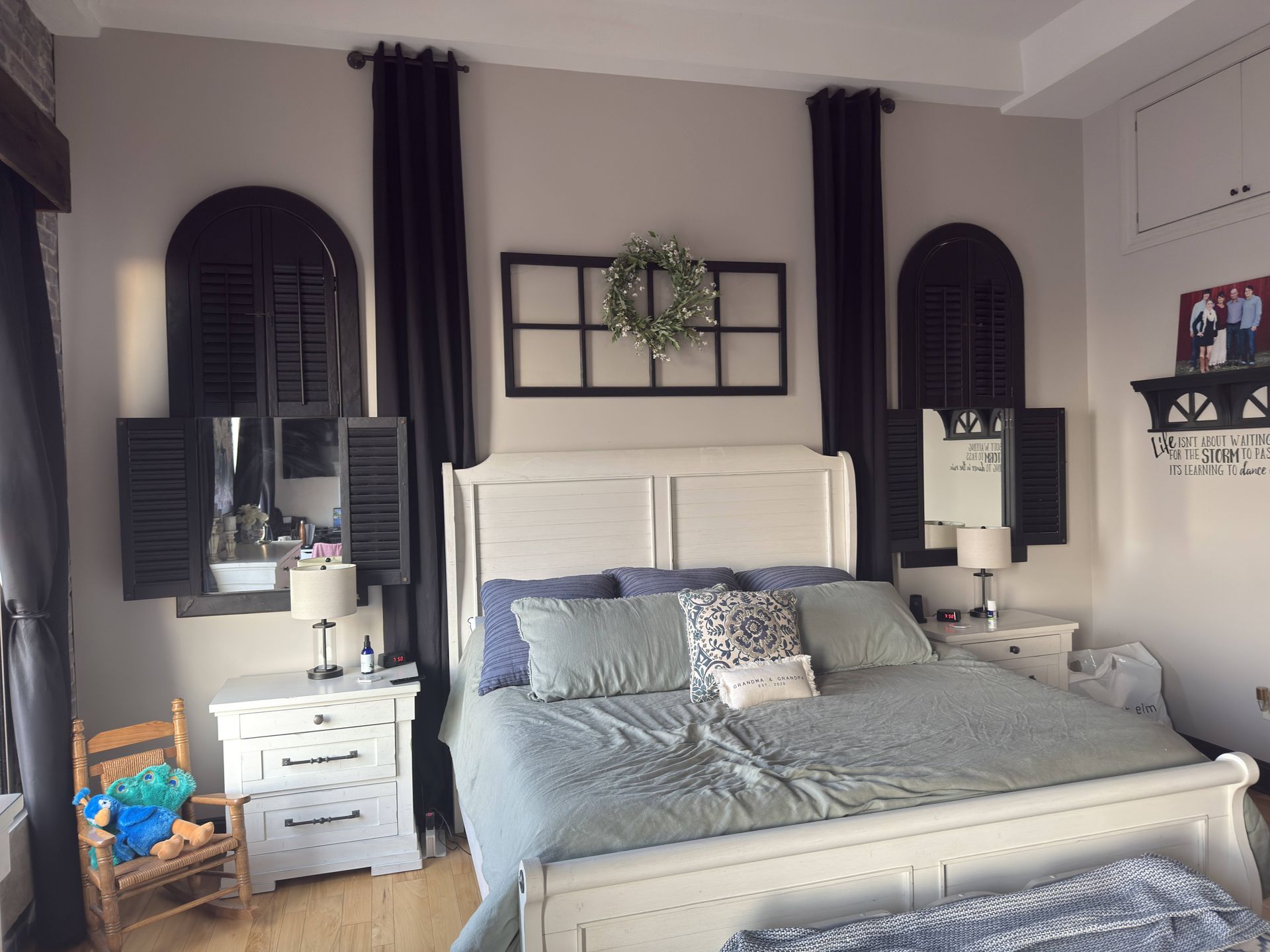 Bedroom with white furniture, arched windows with black shutters, black curtains, and a wreath.