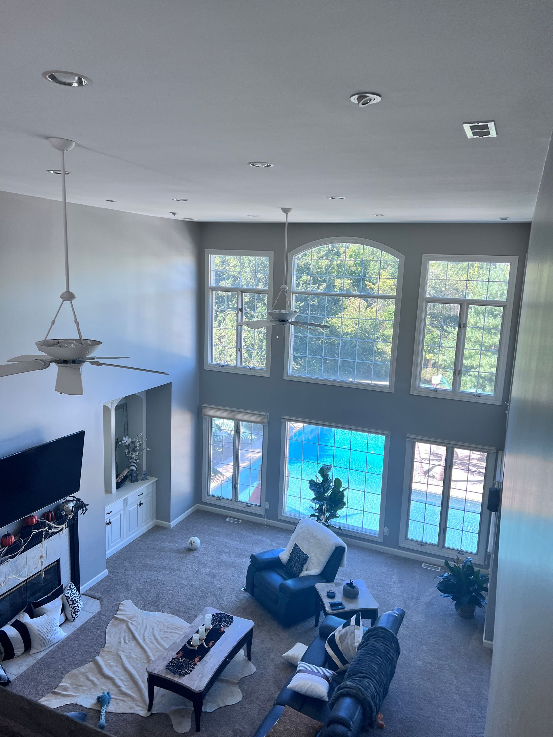 High-ceilinged living room with large windows, fireplace, and seating area in shades of gray, white, and blue.