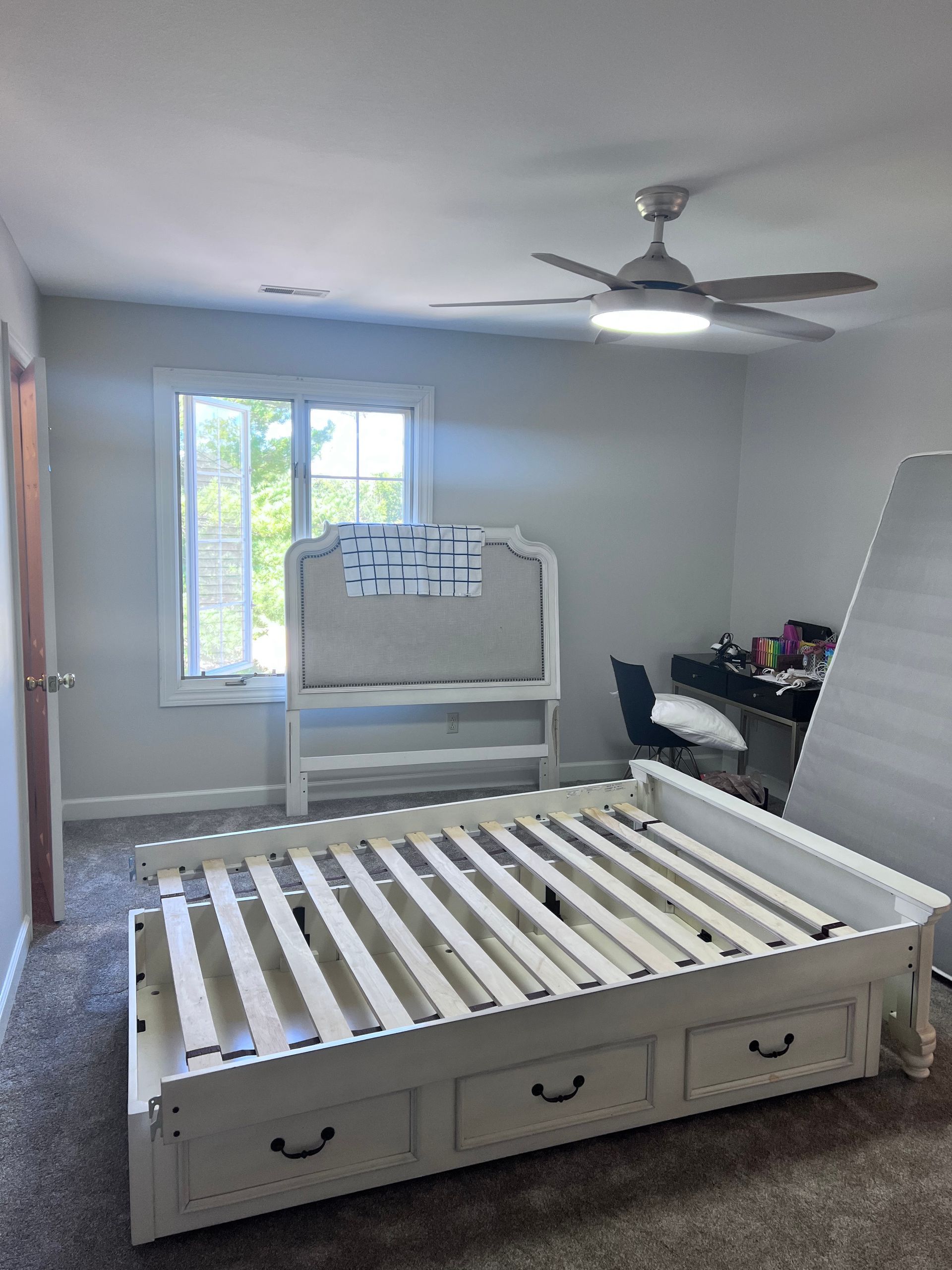 White bed frame with storage drawers and headboard in a bedroom with a window, desk, and ceiling fan.