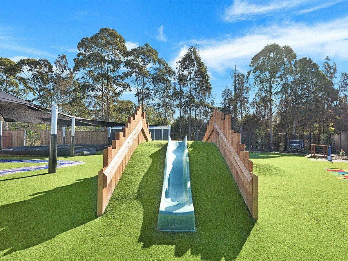 A slide is going down a hill in a playground with trees in the background.