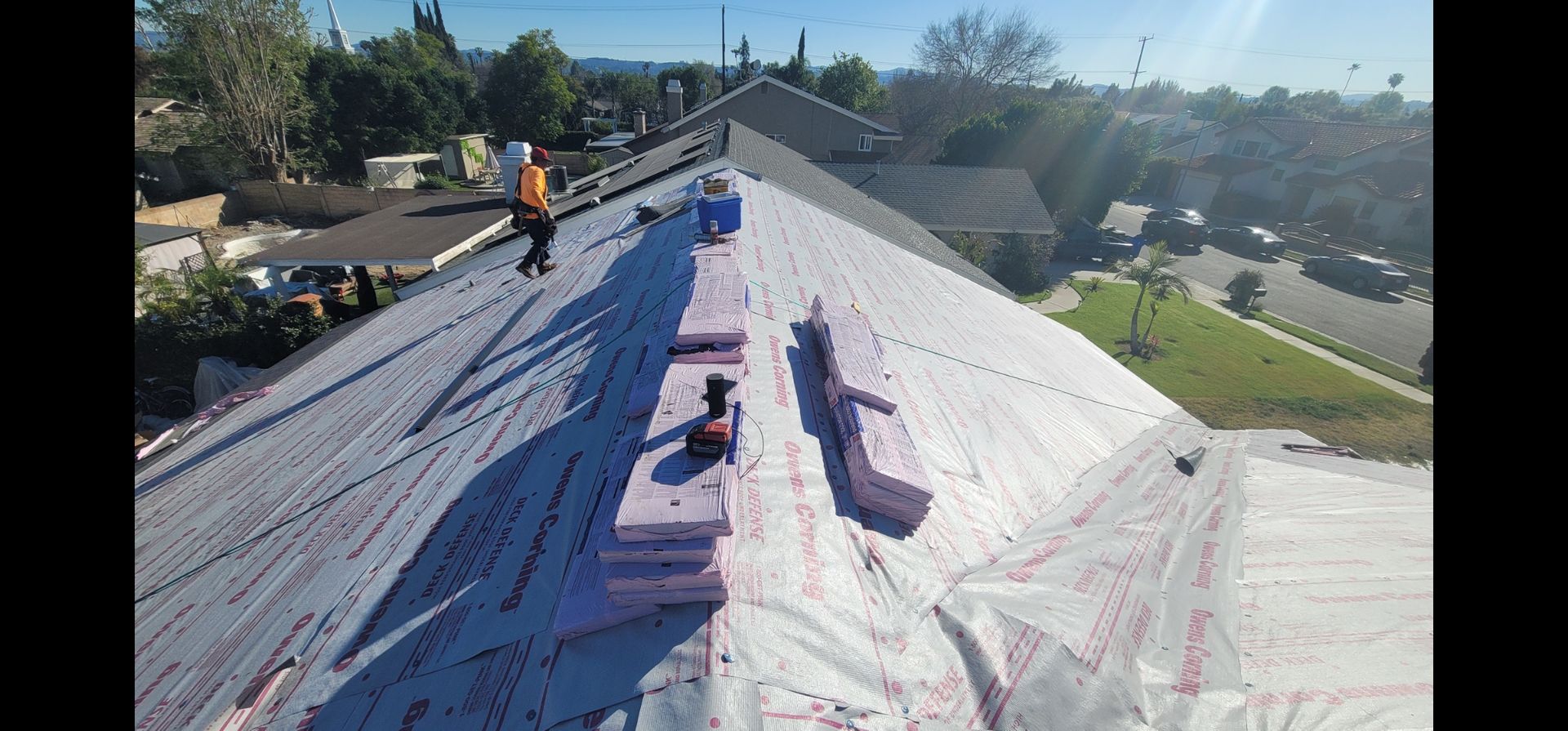 Workers installing roofing materials on a residential roof, with tools and insulation visible. Clear, sunny day.