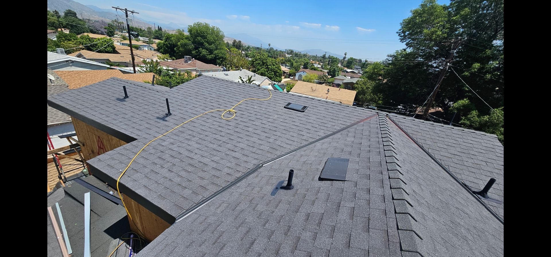 A newly shingled roof with pipes and a skylight, with a clear blue sky and a view of the city.