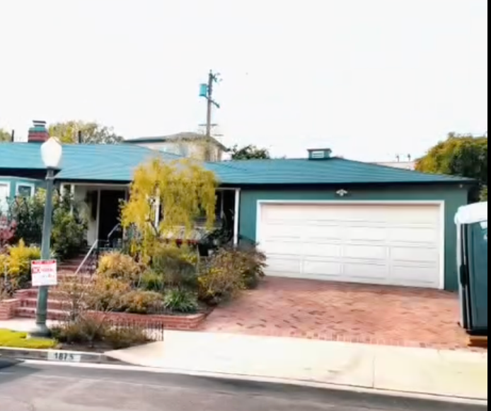 Single-story house with teal roof, white garage door, and brick driveway behind a landscaped front yard
