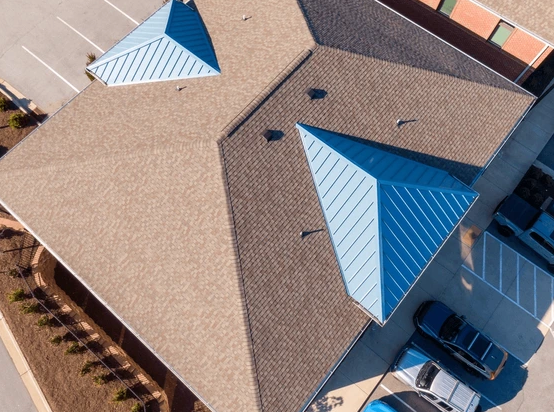 Overhead view of a building's roof with tan shingles and two blue metal accents; parking lot visible.