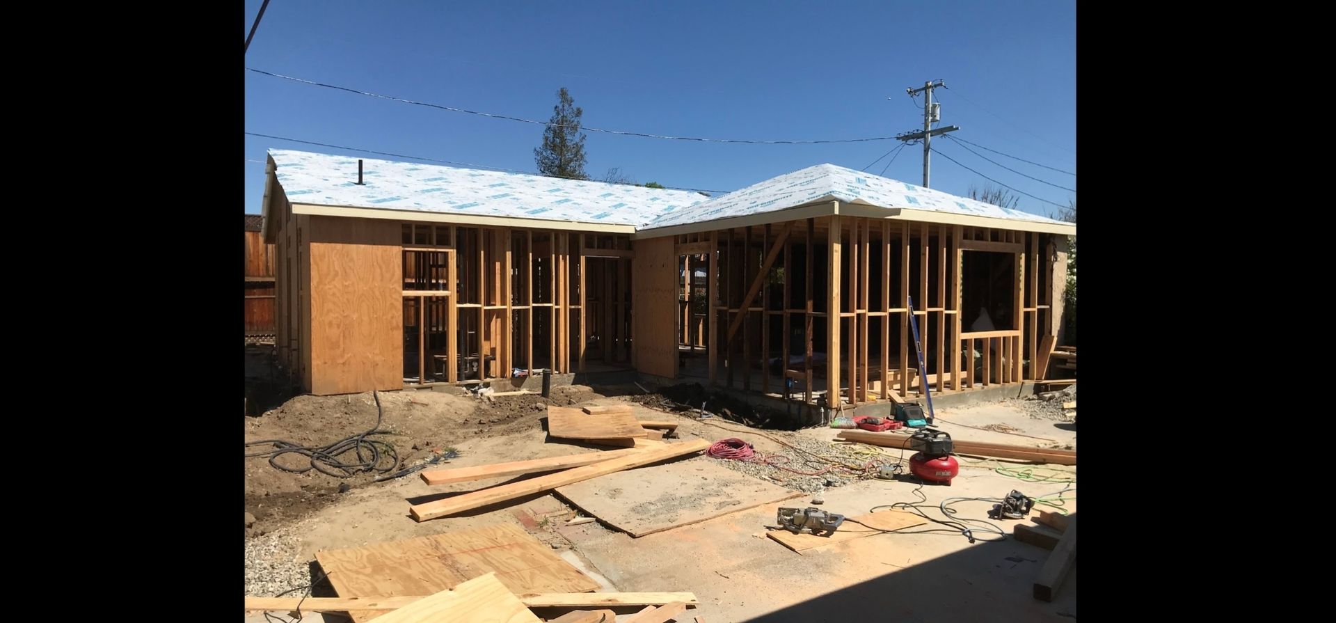 House under construction; wooden frame visible. Roof partially covered, materials scattered. Bright sunny day.