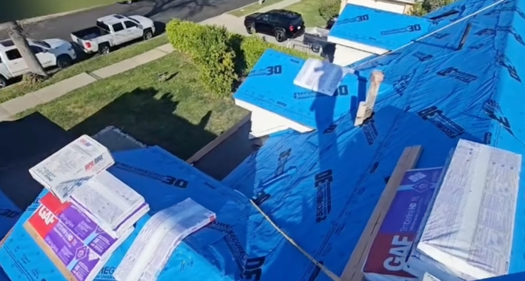 Roof under construction covered in blue tarp and plywood, seen from above with parked cars nearby