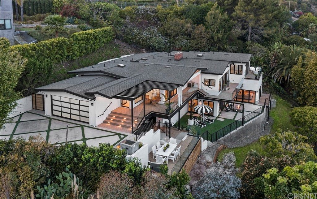 Aerial view of a white multi-level house with a dark roof and patio, surrounded by green foliage.