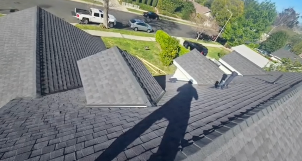 Rooftop view of dark gray shingles with a worker’s shadow and neighboring houses below.
