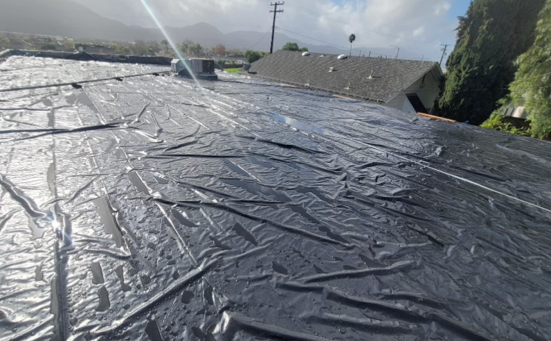 Black plastic sheeting covering a rooftop, with mountains and houses in the background under a cloudy sky