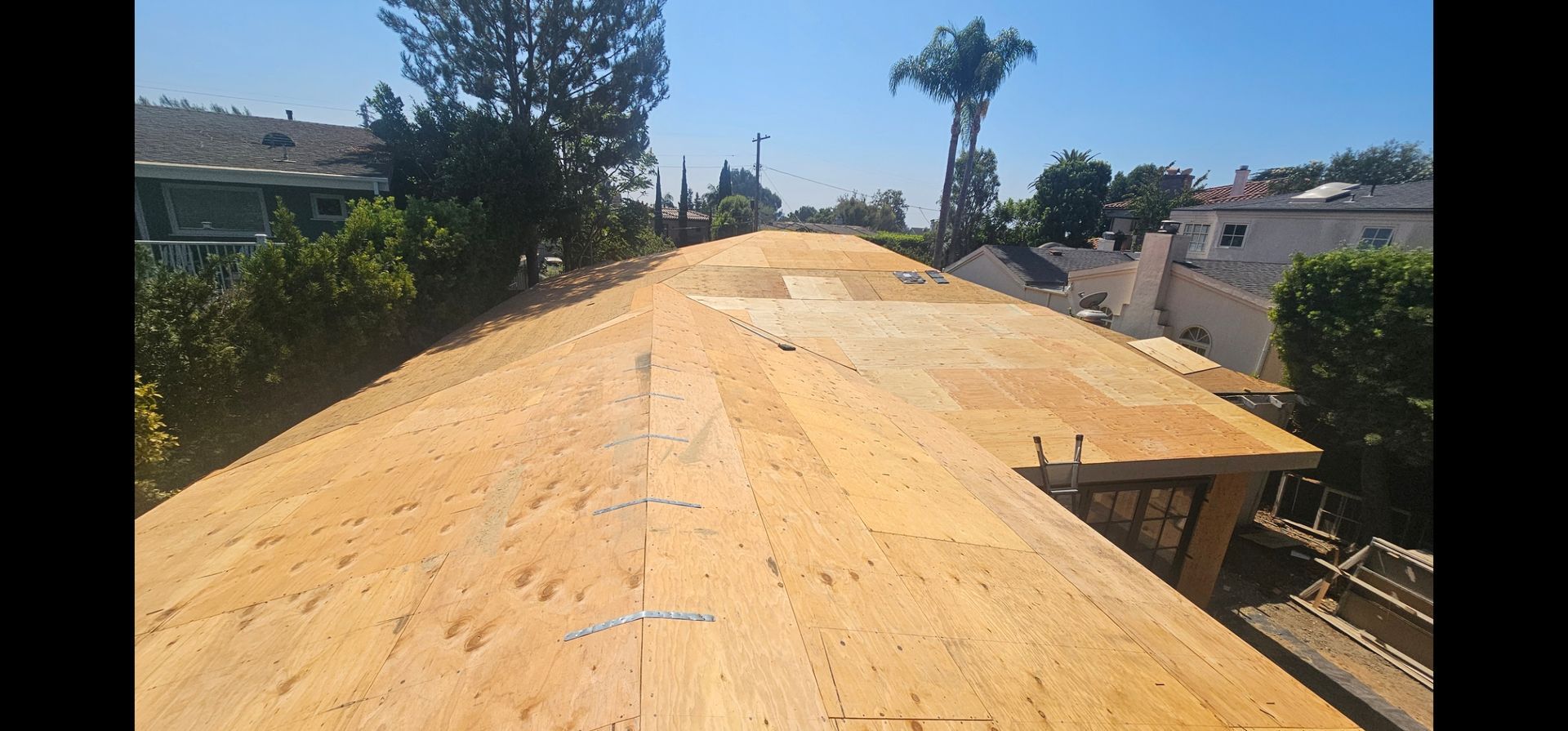 View of a roof being built, wooden planks are laid out. Blue sky and surrounding homes visible.