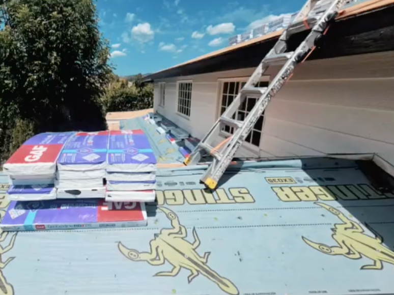 Roofers installing insulation on a house roof under a blue sky, with ladders and stacked materials.