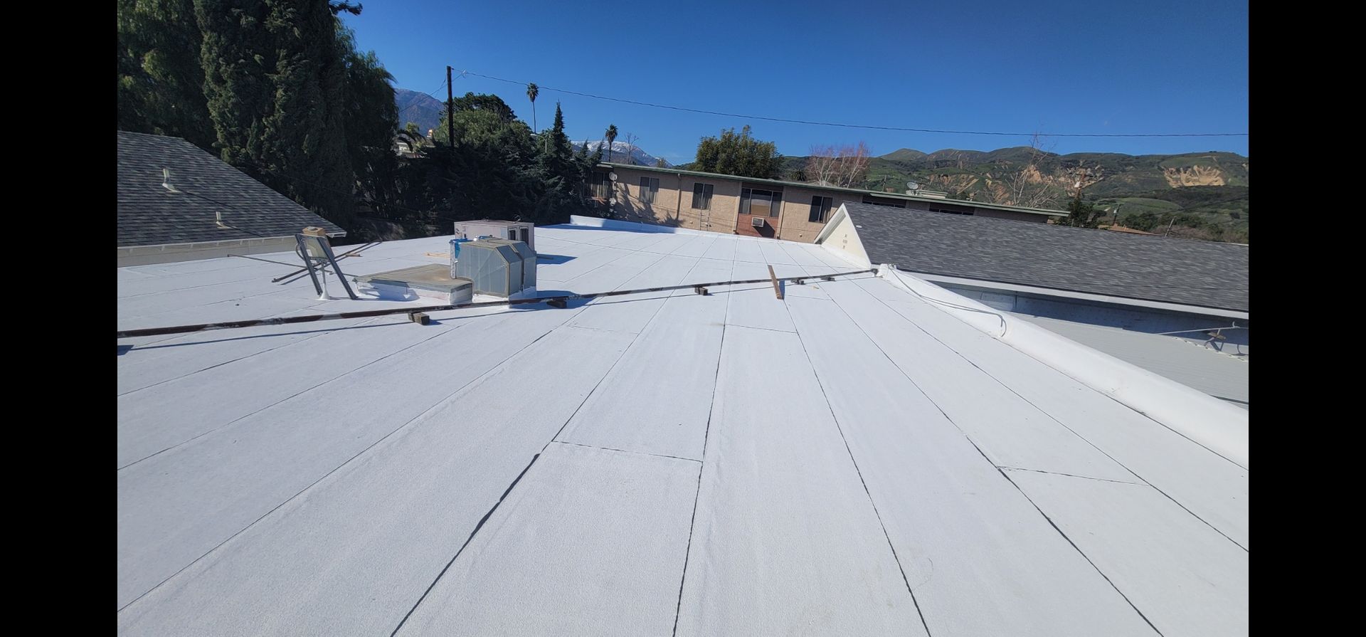 White roof with ventilation unit, against a clear blue sky. Buildings and trees in background.
