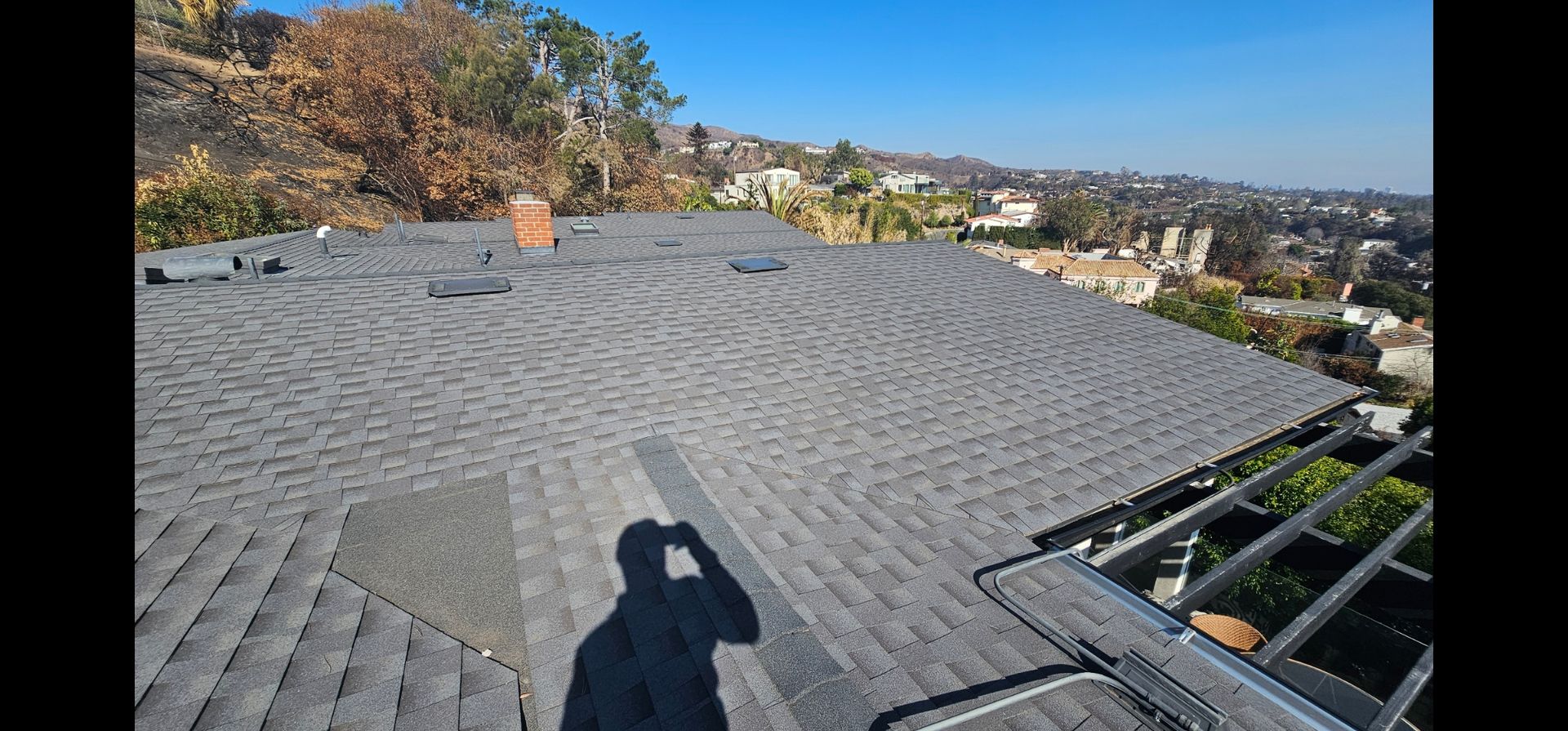 View from rooftop with grey shingles overlooking a suburban landscape under a clear blue sky.