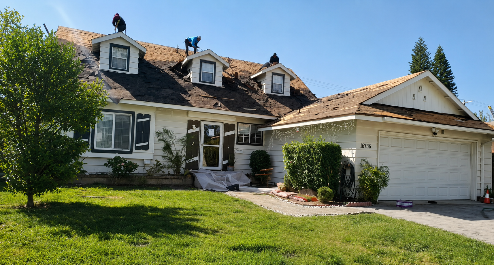 Suburban house with beige siding, brown roof, white garage, and green lawn on a sunny day