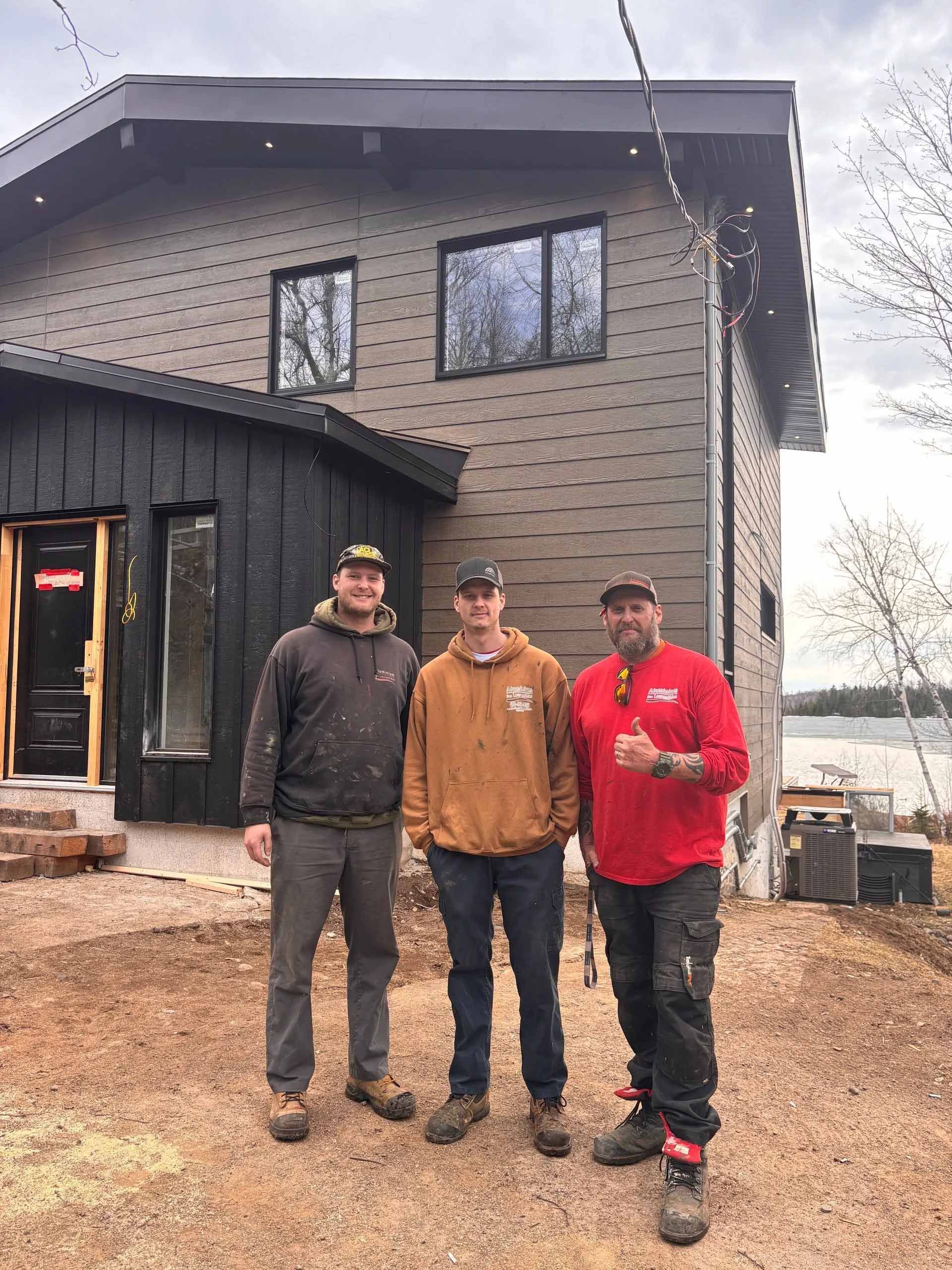 Trois personnes sourient devant une maison en construction, avec un bardage en bois sombre et un premier plan recouvert de gravier.