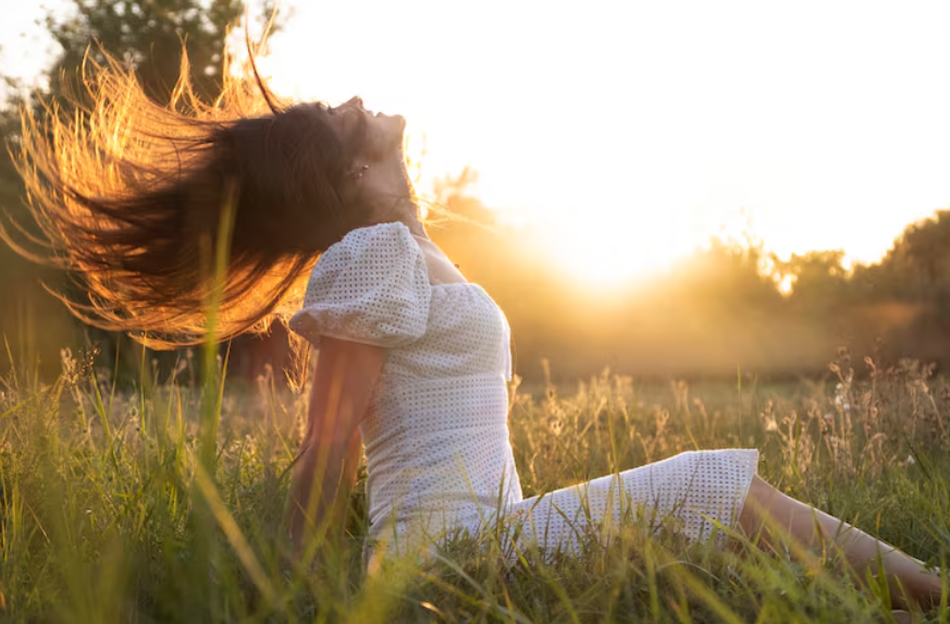 Mujer con vestido blanco, con la cabeza hacia atrás y el cabello fluyendo en un campo iluminado por el sol.