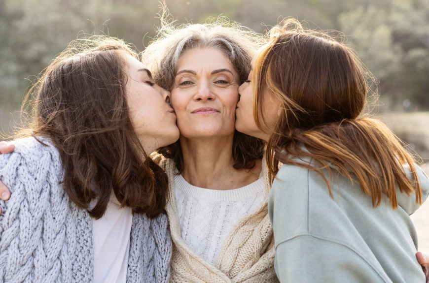 Dos mujeres jóvenes besando a una mujer mayor en las mejillas; al aire libre, soleado.