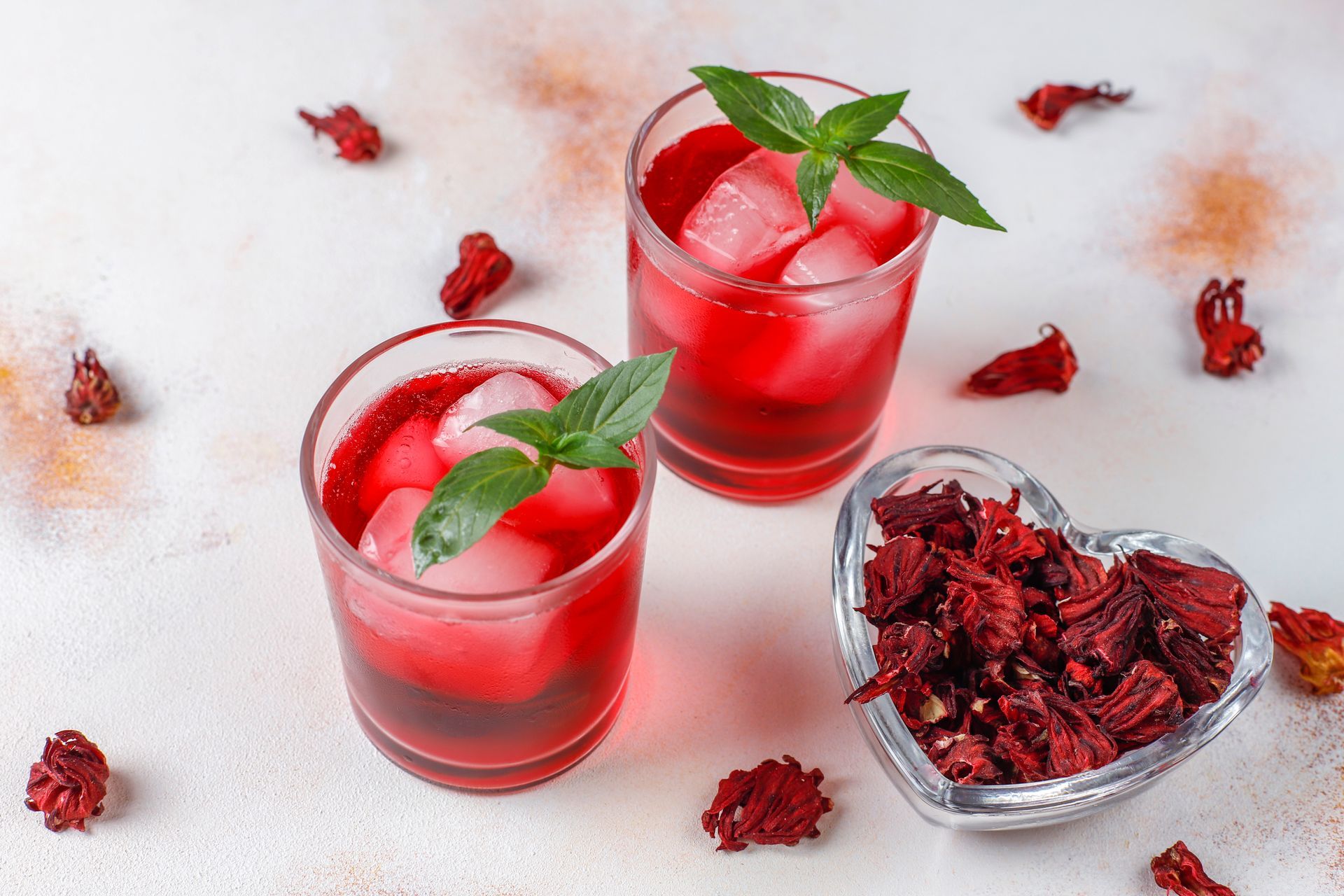 Two glasses of hibiscus tea with ice cubes and a heart shaped bowl of dried hibiscus flowers.