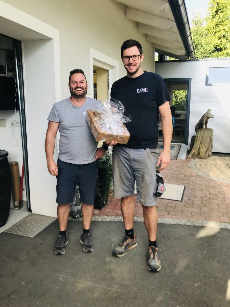 Zwei Männer stehen nebeneinander vor einem Haus und halten eine Kiste.